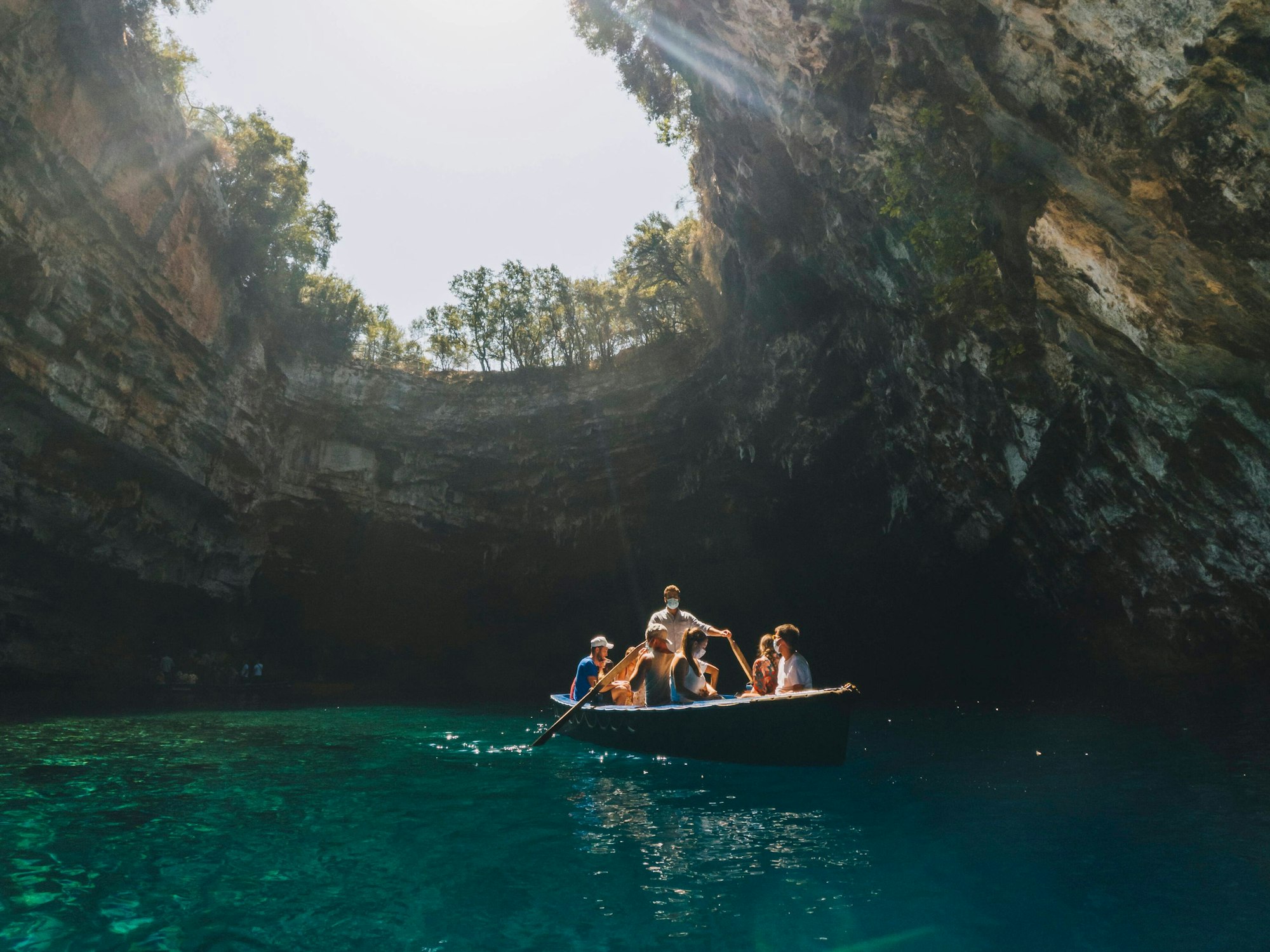 Ein Boot mit Touristinnen und Touristen fährt durch den blauen See der Melissani Höhle. Oben im Bild sieht man die Öffnung der Höhle und den Himmel.