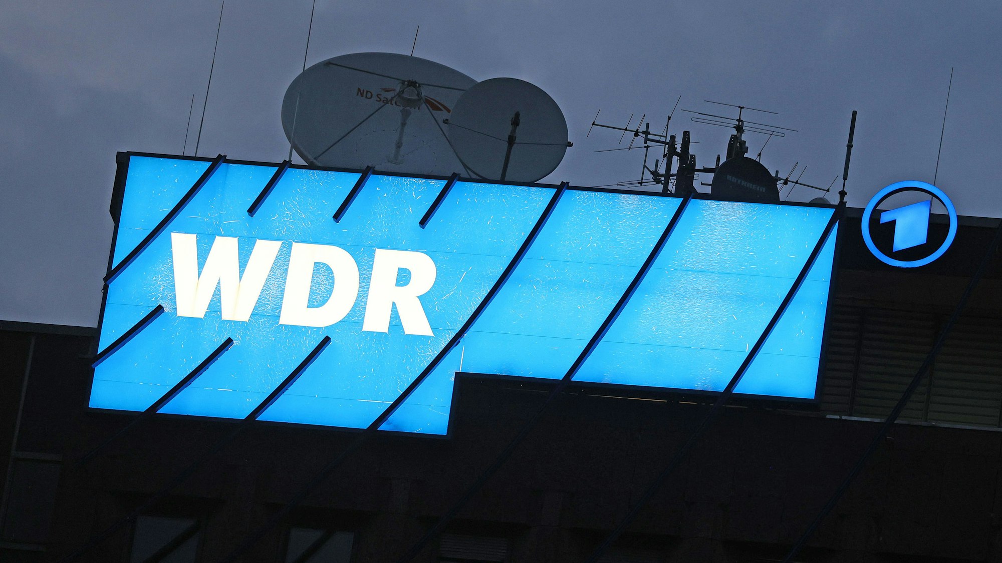 Das Logo des Nachrichtensenders WDR leuchtet auf dem Dach des WDR-Gebäudes in der Kölner Innenstadt.