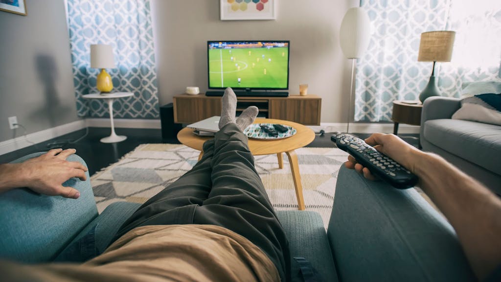 Auf dem Foto sitzt ein Mann auf einem Sessel und schaut Fußball auf einem kleinen Fernseher.