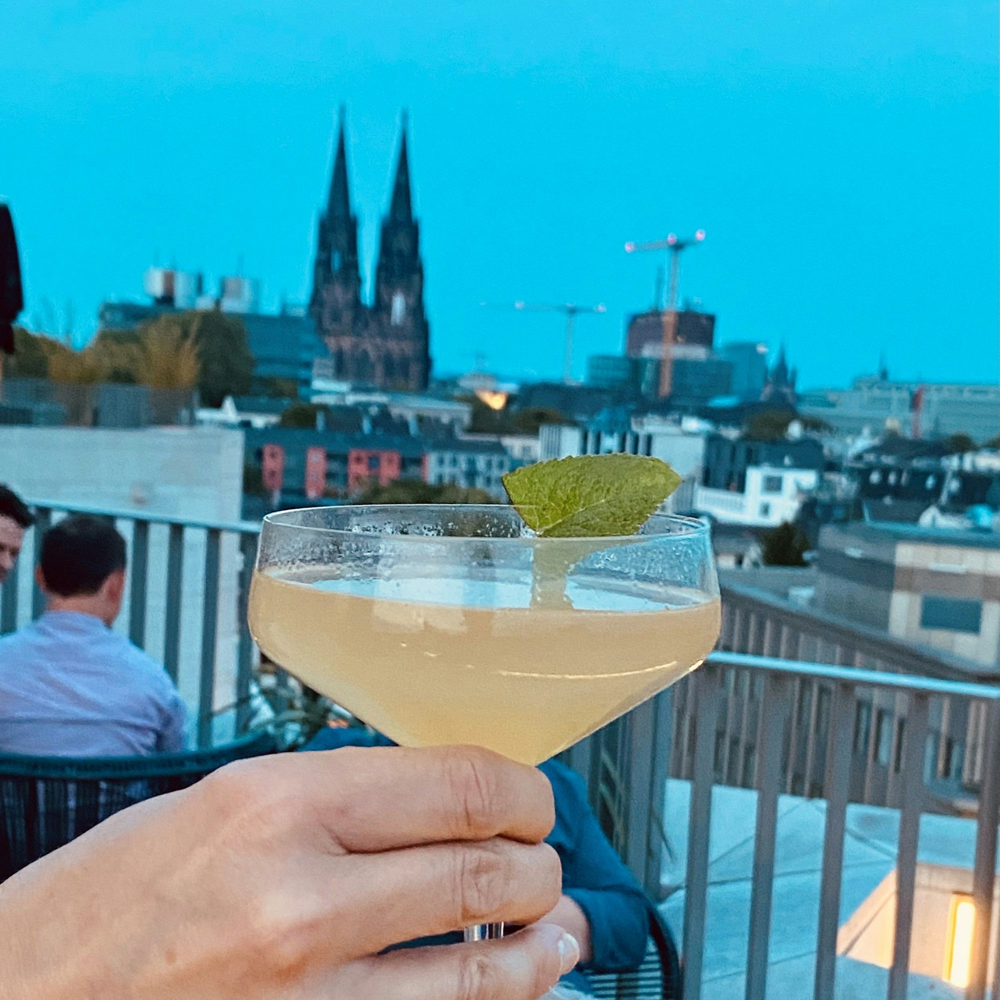 Cocktail im Vordergrund, der Kölner Dom und Köln-Panorama bei Abendlicht im Hintergrund