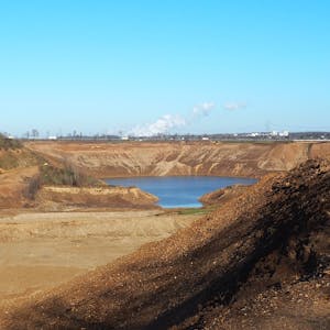 Hinter Hügeln aus braunem Sand und Kies ist eine große Fläche Wasser zu sehen, das sich in der Kiesgrube ansammelt. Links am Rand der Grube arbeiten Bagger.