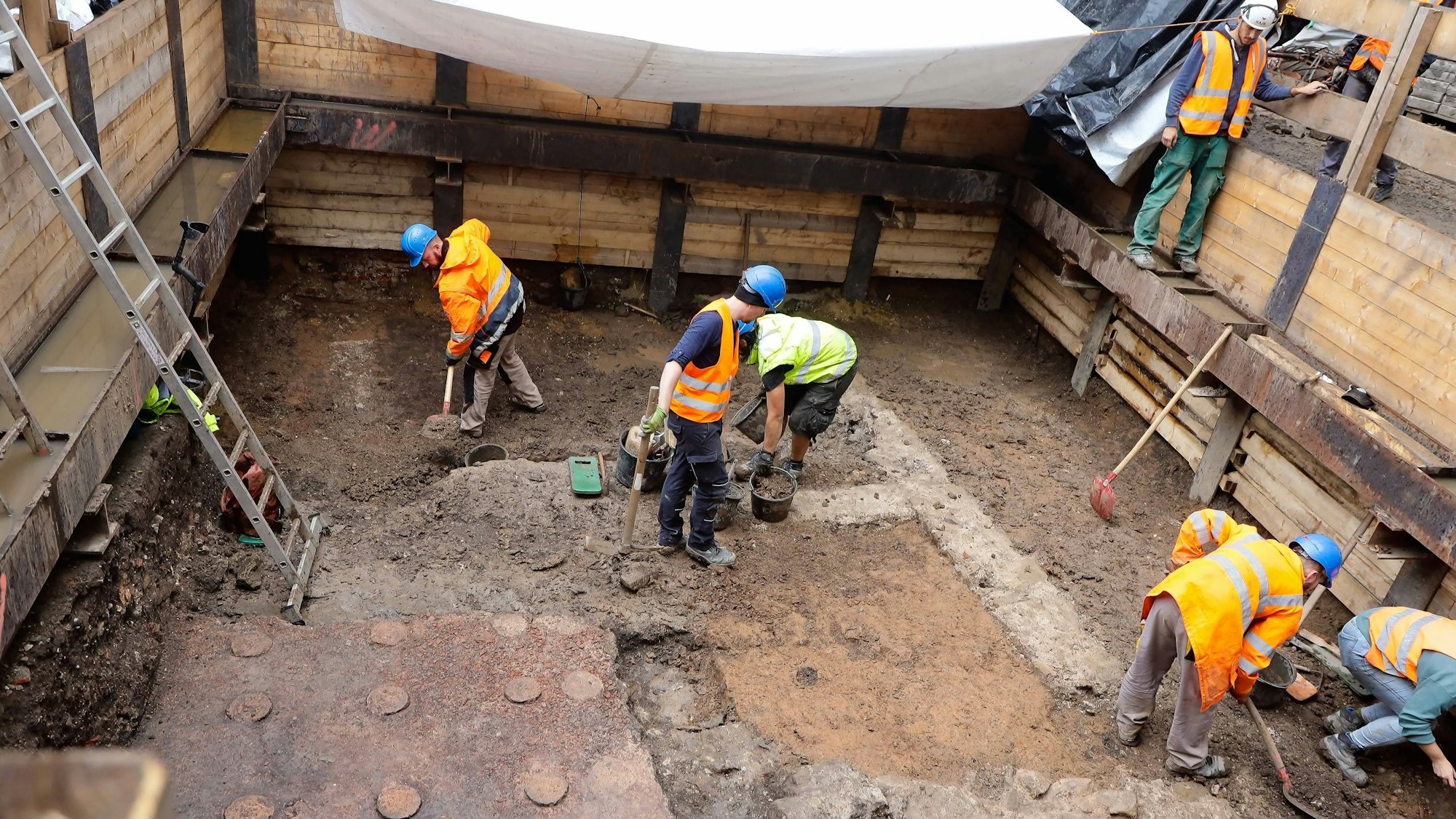 Zu sehen sind mehrere Bauarbeiter in orangefarbenen Warnwesten, die mit der Ausgrabung einer römischen Badeanlage am Kölner Neumarkt beschäftigt sind.