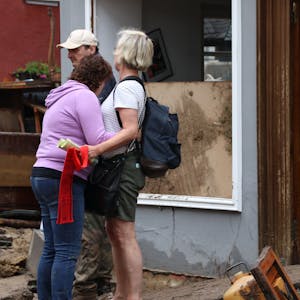 Das Bild zeigt zwei Frauen, die sich nach der Flutkatastrophe in Bad Münstereifel trösten. Im Hintergrund sind ein Mann und ein zerstörtes Haus zu sehen.