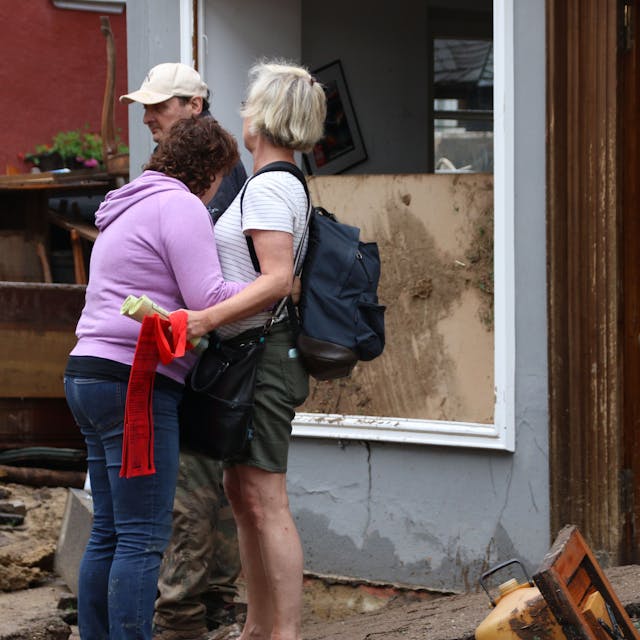 Das Bild zeigt zwei Frauen, die sich nach der Flutkatastrophe in Bad Münstereifel trösten. Im Hintergrund sind ein Mann und ein zerstörtes Haus zu sehen.