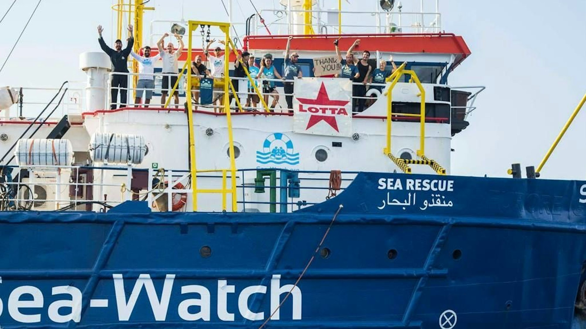Sea-Watch Crew Mitglieder auf dem grüßen mit dem Lotta Plakat vom Boot.