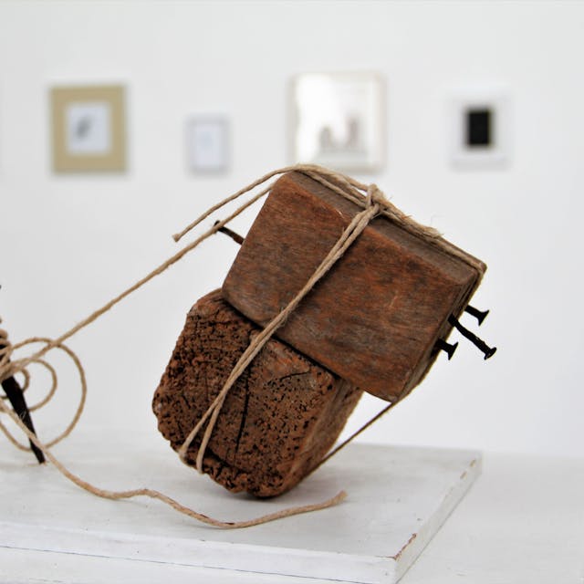 Ein Holzobjekt, das mit Bindfäden an einem Nagel befestigt ist, steht in einem Ausstellungsraum.