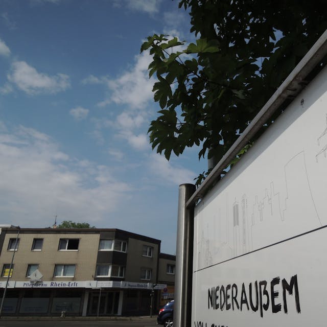 Auf einem Schild aus Metall steht „Niederaußem“, im Hintergrund sind Gebäude.