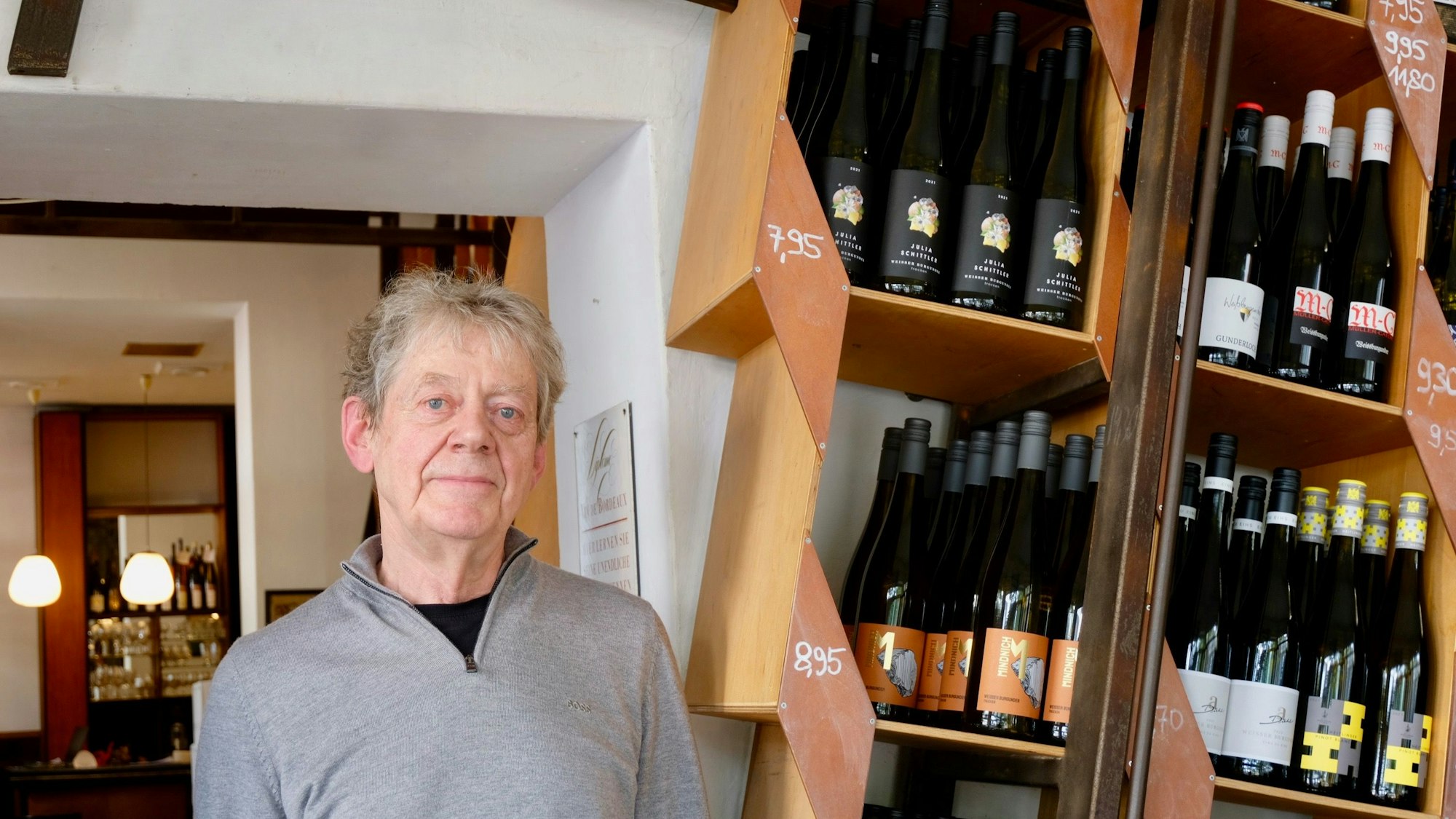 Ein älterer Mann steht in einem Weinladen, neben ihm sind Regale mit Weinflaschen zu sehen.
