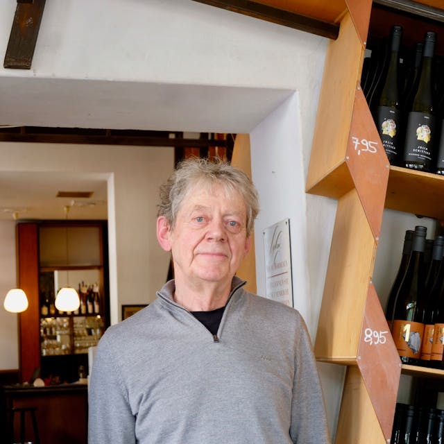 Ein älterer Mann steht in einem Weinladen, neben ihm sind Regale mit Weinflaschen zu sehen.