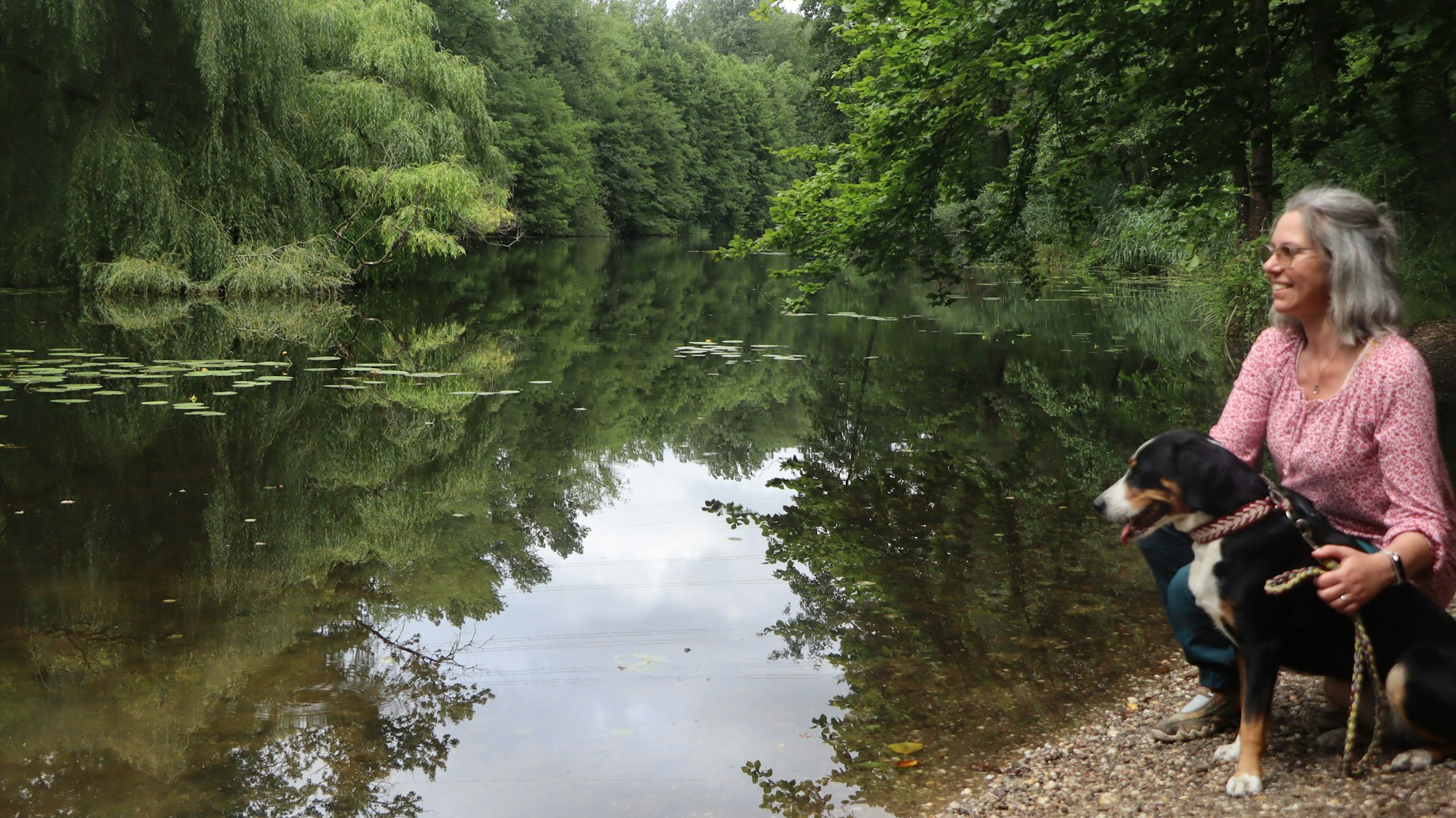 Eine Frau mit Hund kniet am Ufer eines Sees in einem Wald.