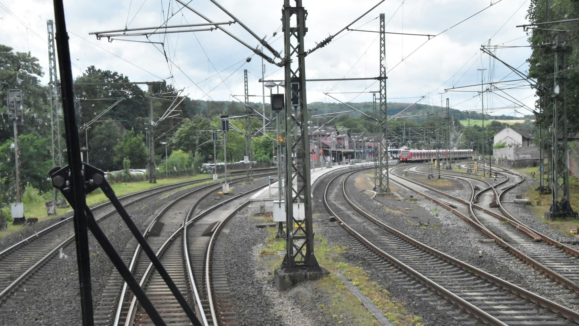 Blick aus dem Führerstand eines Regionalexpresszugs. Zu sehen sind die Schienen, Oberleitungsmasten und ein rotes Bahnhofsgebäude im Hintergrund. Auf einem Gleis stehen rote Waggons einer S-Bahn.