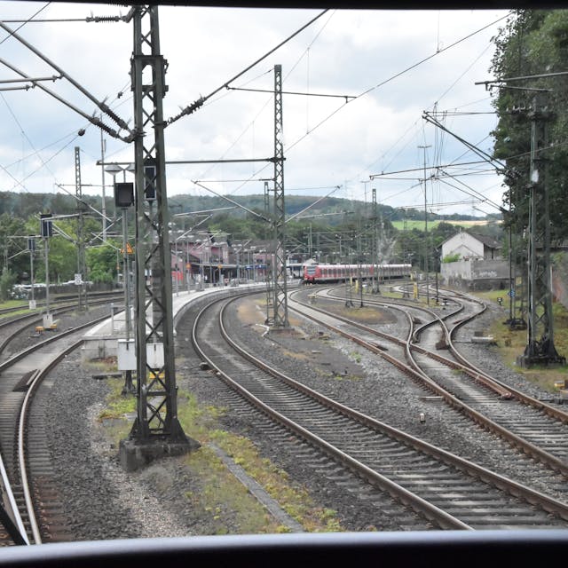 Blick aus dem Führerstand eines Regionalexpresszugs. Zu sehen sind die Schienen, Oberleitungsmasten und ein rotes Bahnhofsgebäude im Hintergrund. Auf einem Gleis stehen rote Waggons einer S-Bahn.