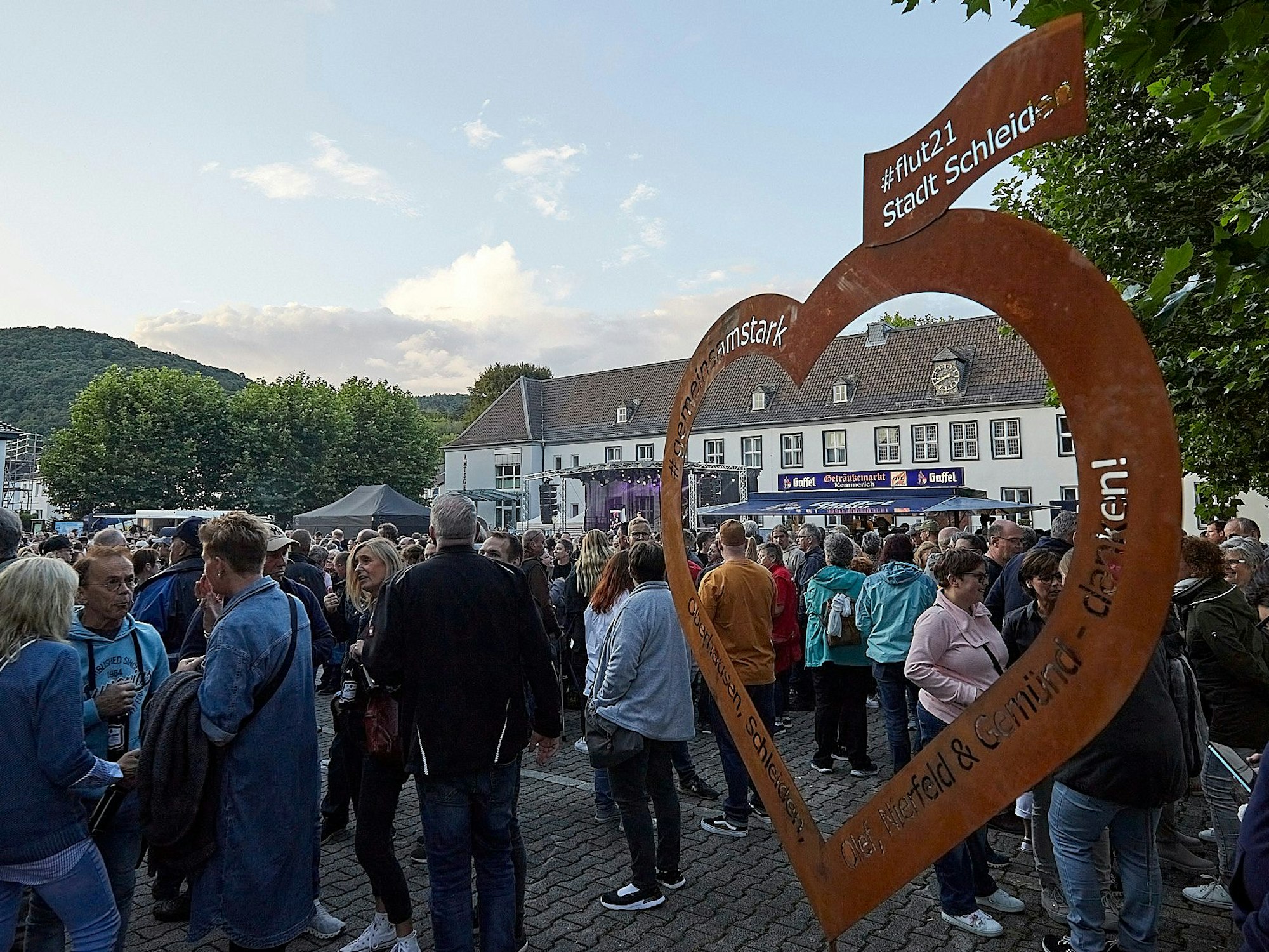 Das Bild zeigt ein Herz aus rostigem Blech, auf dem ein Dank an die Flut-Helfer steht. Im Hintergrund sind zahlreiche Menschen auf dem Marienplatz zu sehen.