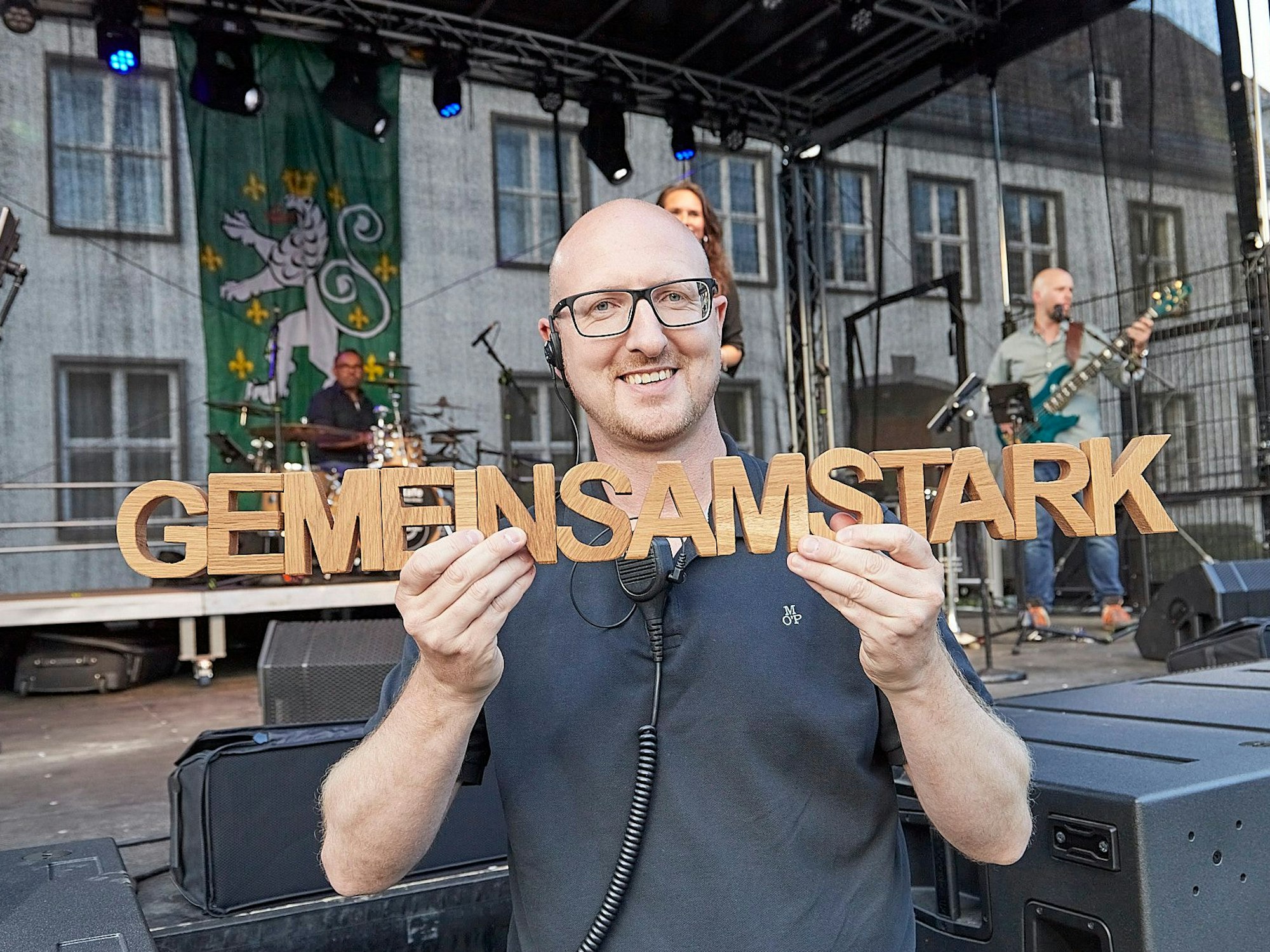 Schleidens Bürgermeister Ingo Pfennings hält einen aus Holz gefertigten Schriftzug „gemeinsam stark“ in beiden Händen. Im Hintergrund ist eine Bühne zu sehen.