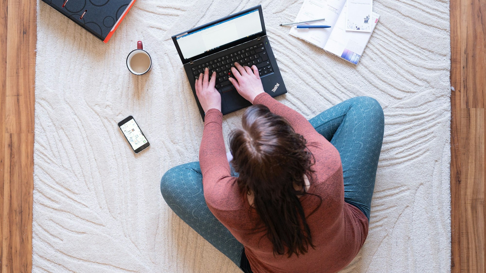 Eine Frau sitzt mit Laptop, Kaffeetasse, ihrem Handy und einem Ordner auf einem Teppich und arbeitet.