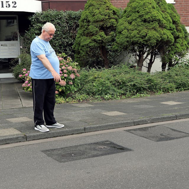 Ein Mann steht vor einem Mehrfamilienhaus an der Straße und zeigt auf geflickten Asphalt.