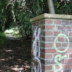 Das Bild zeigt ein mit Graffiti beschmiertes Backsteintor und dahinter einen Waldweg.