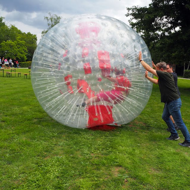 Zwei Männer rollen einen riesigen Plastikball, in dem eine Frau versucht, zu laufen.