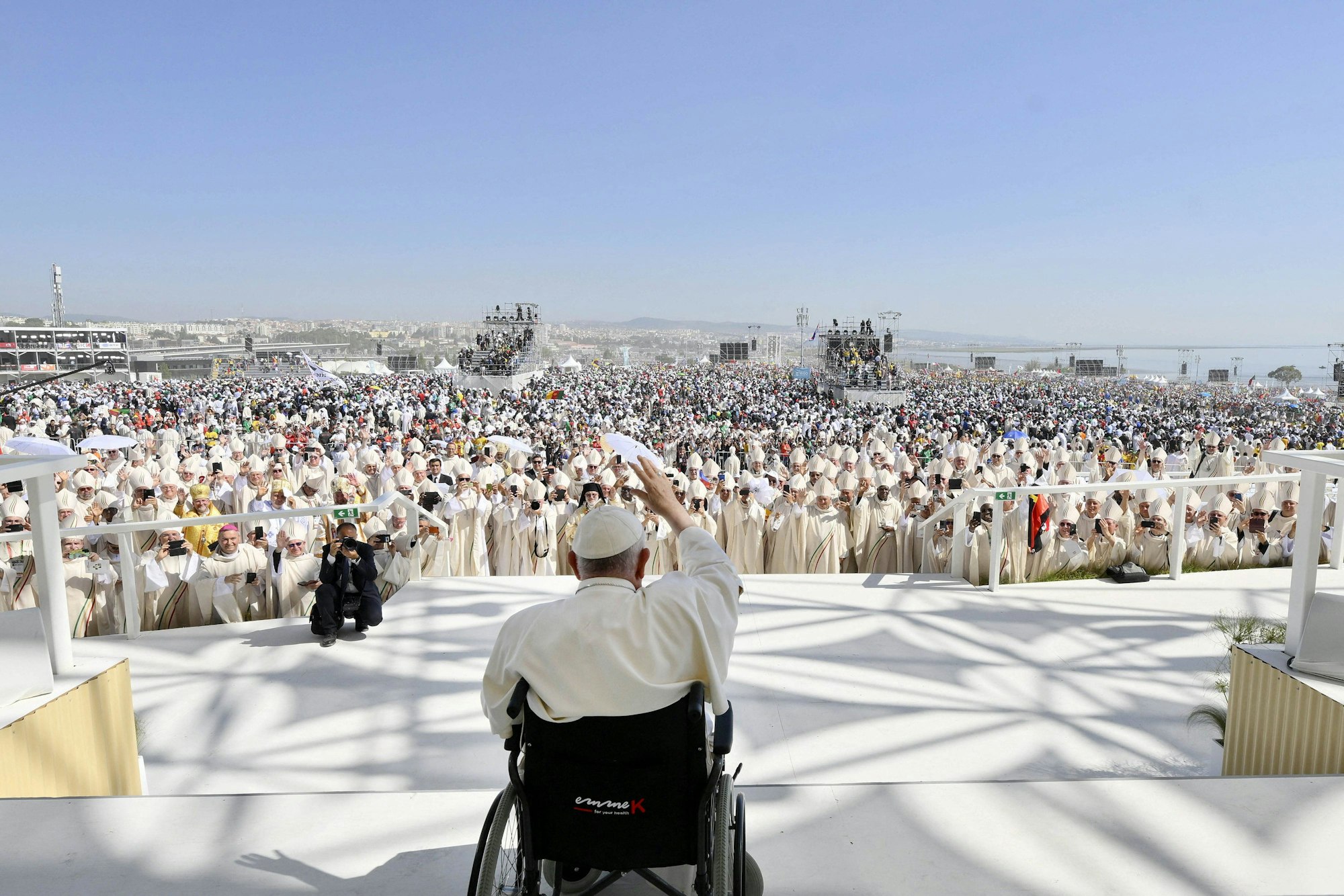 Papst Franziskus sitzt im Rollstuhl auf einer Bühne unter freiem Himmel. Das Bild zeigt ihn von hinten. Im Hintergrund sieht man die Priester und Gäubigen vor der Bühne stehen.