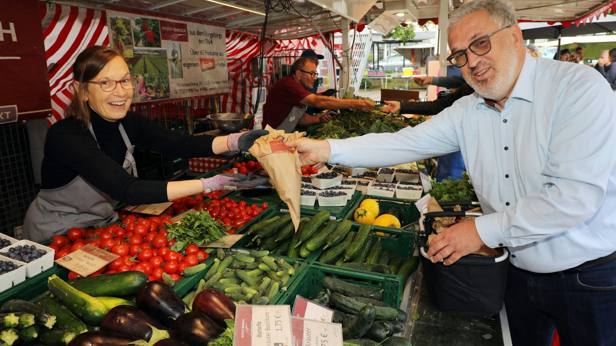 Eine Frau an einem Marktstand reicht einem Mann eine Papiertüte über die Auslagen des Obst- und Gemüsestands auf dem Hoffnungsthaler Wochenmarkt.