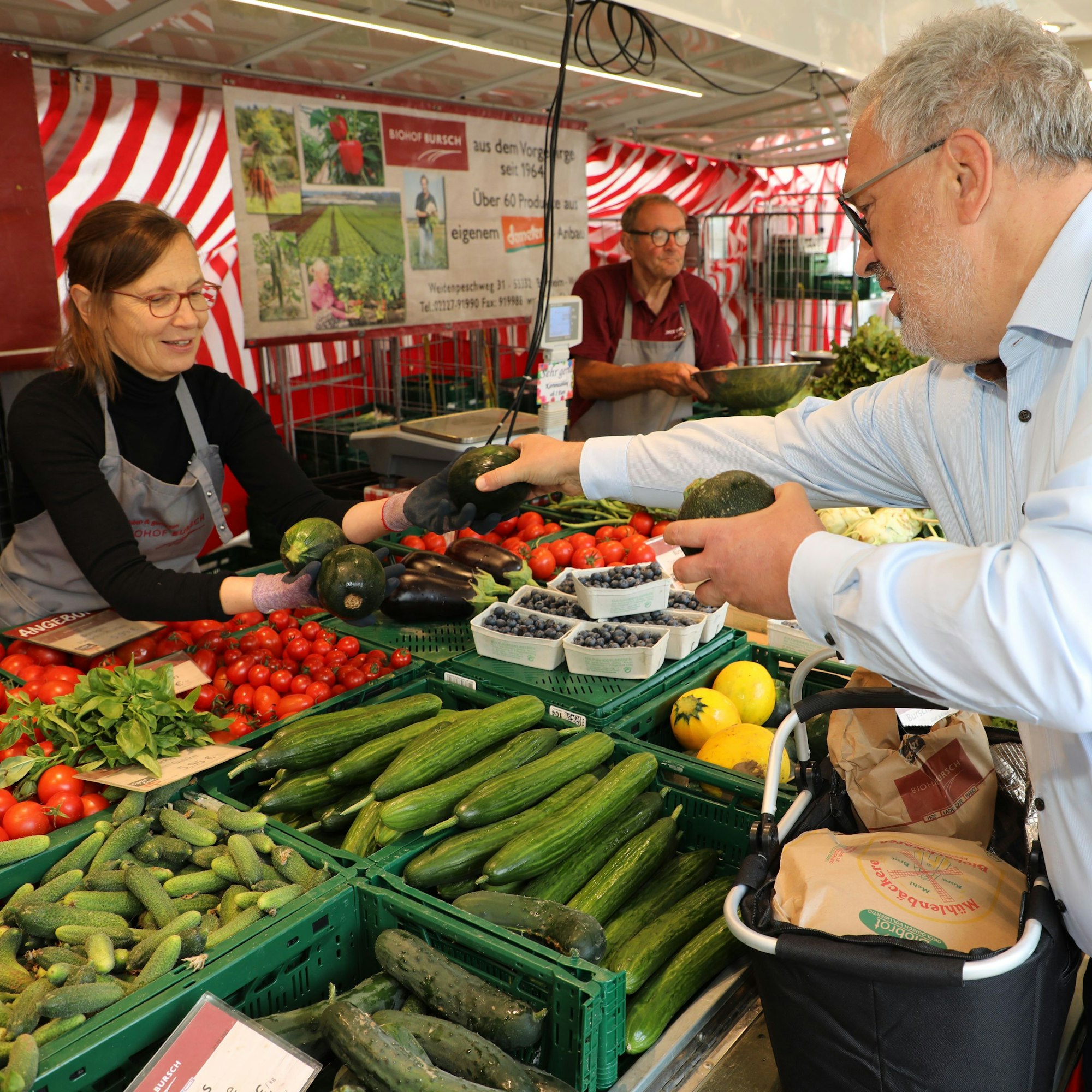 Gerhard Zorn reicht Gemüse über die Auslagen des Standes des Biohofs Busch auf dem Hoffnungsthaler Wochenmarkt an die Verkäuferin.