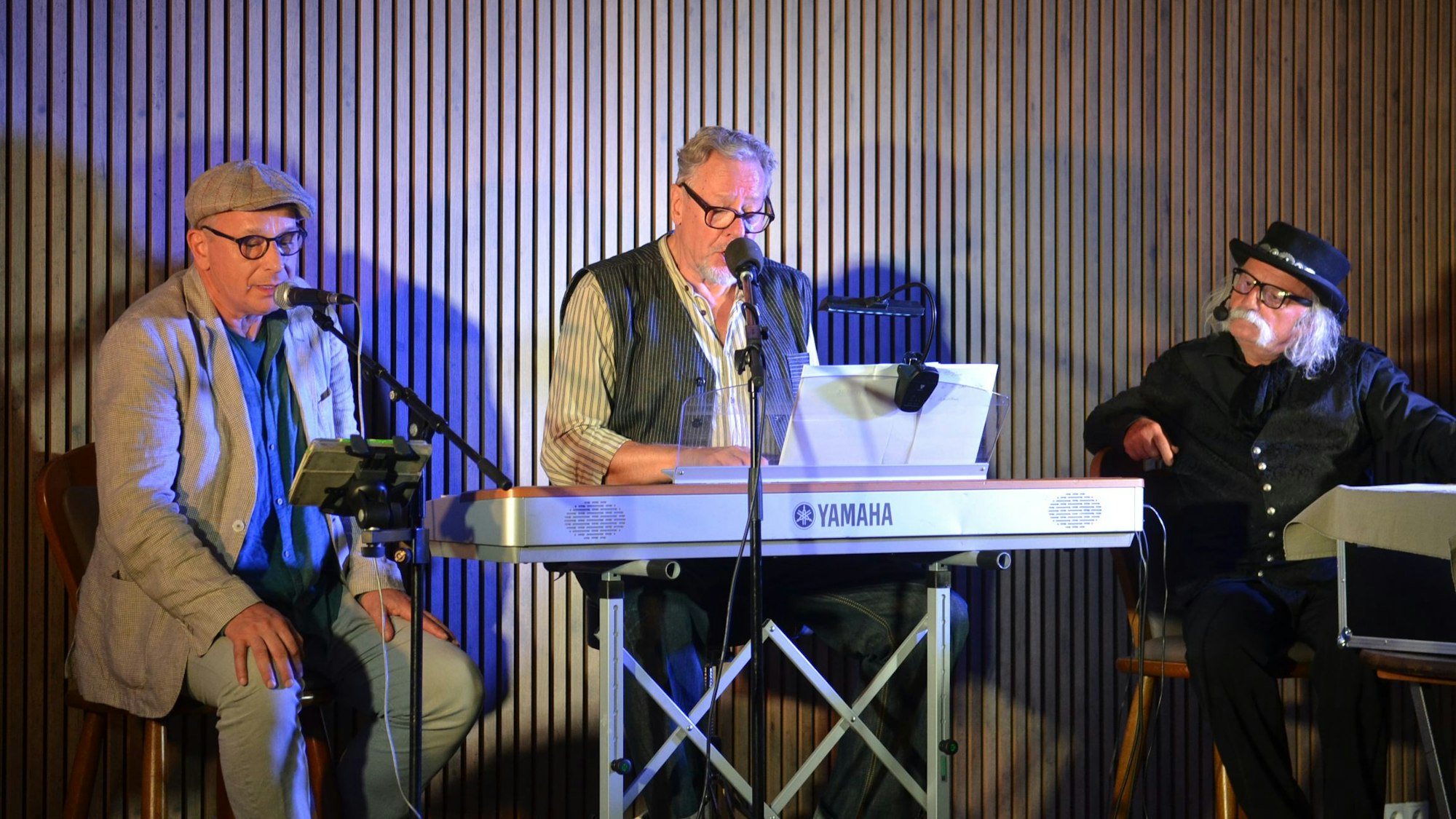 Der Sänger Wolfgang Reyscher sitzt an einem Keyboard, neben ihm sitzen die Kabarettisten Achim Konejung (l.) und Hubert vom Venn (r.).
