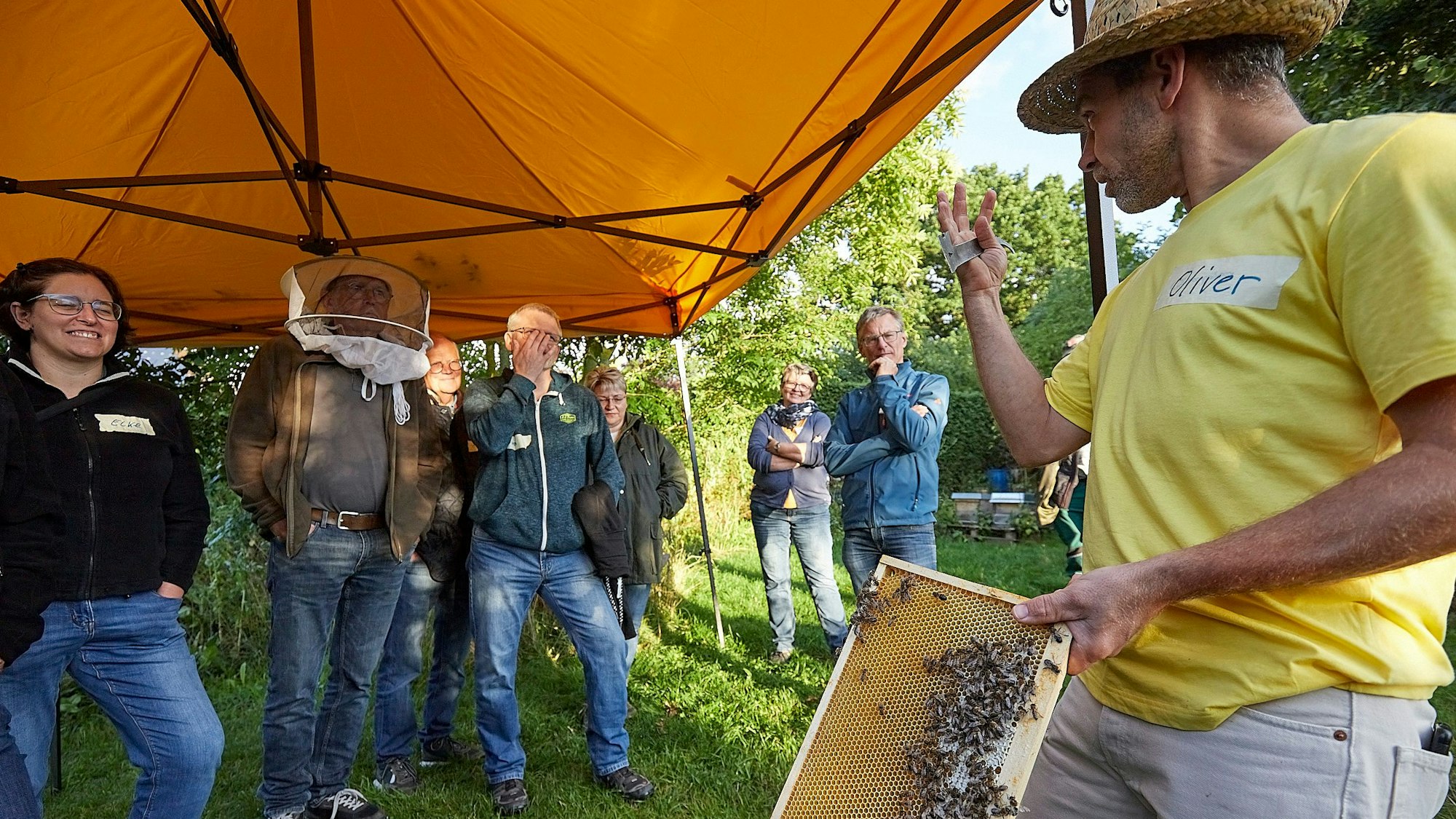 Das Bild zeigt Oliver Tretbar (r.). Er hält eine Bienenwabe in der Hand und erklärt einer Gruppe von Menschen etwas.