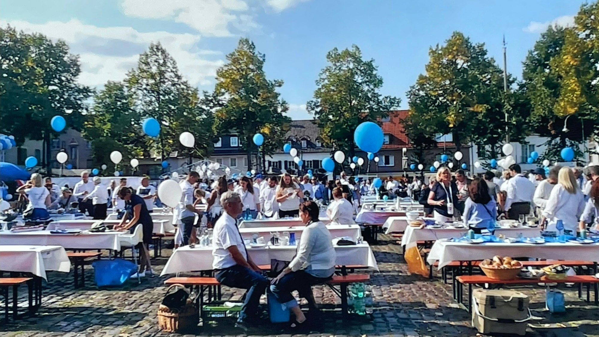 Viele geschmückte Biertische stehen auf einem Marktplatz, Menschen stehen und sitzen um sie herum. Luftballons in Blau und Weiß sind zu sehen.