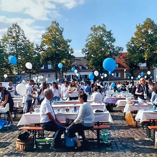 Viele geschmückte Biertische stehen auf einem Marktplatz, Menschen stehen und sitzen um sie herum. Luftballons in Blau und Weiß sind zu sehen.