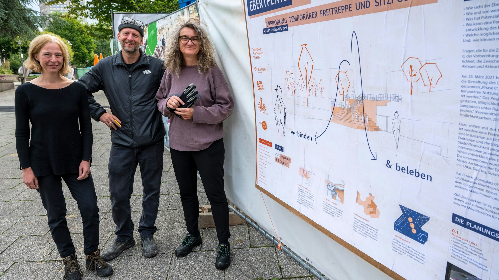 Susanne Kohte, Chris Schroeer-Heiermann und Yasemin Utku stehen an der Baustelle am Ebertplatz vor dem Projektbanner.