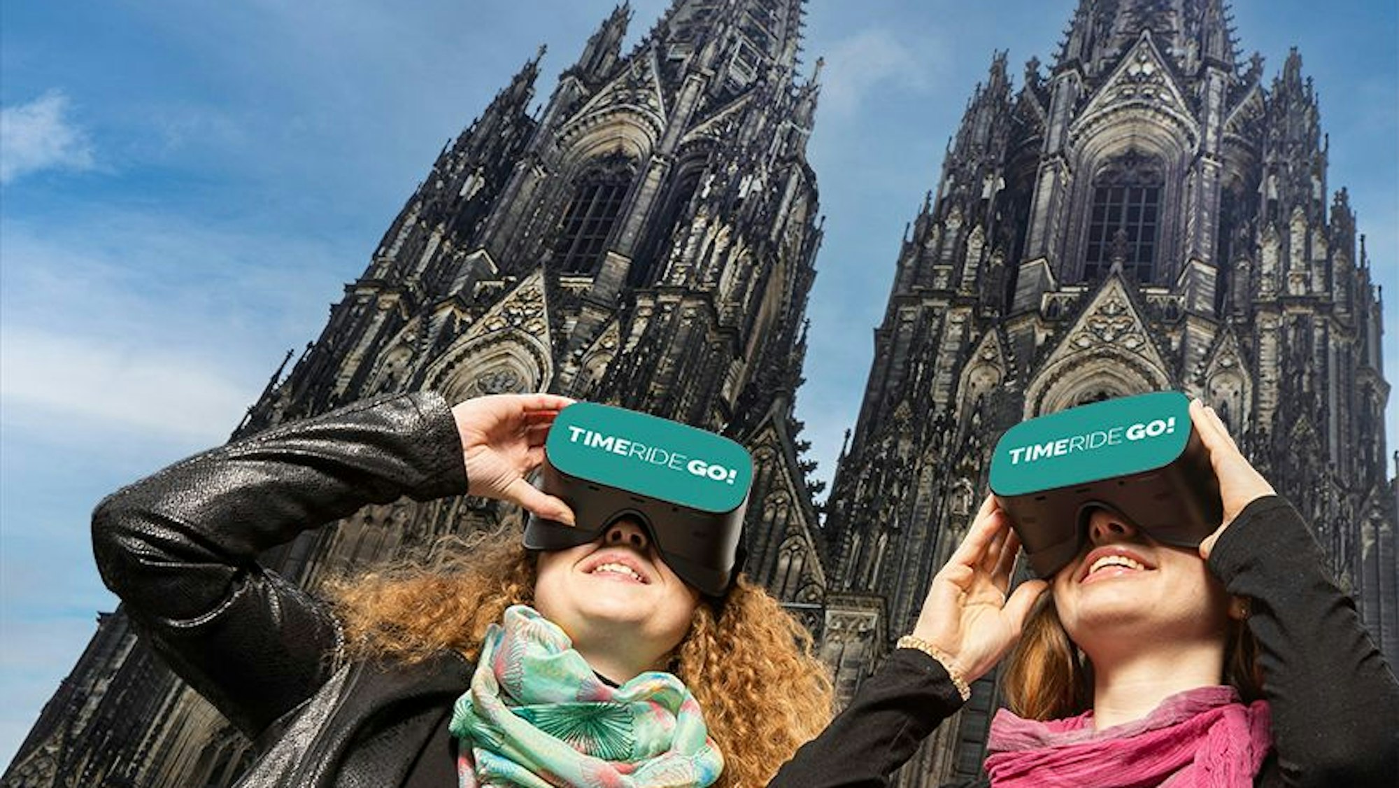 Zwei Frauen stehen vor dem Kölner Dom und schauen durch VR-Brillen mit der Aufschrift „Time Ride Go“
