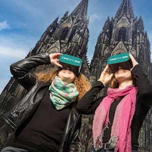 Zwei Frauen stehen vor dem Kölner Dom und schauen durch VR-Brillen mit der Aufschrift „Time Ride Go“&nbsp;