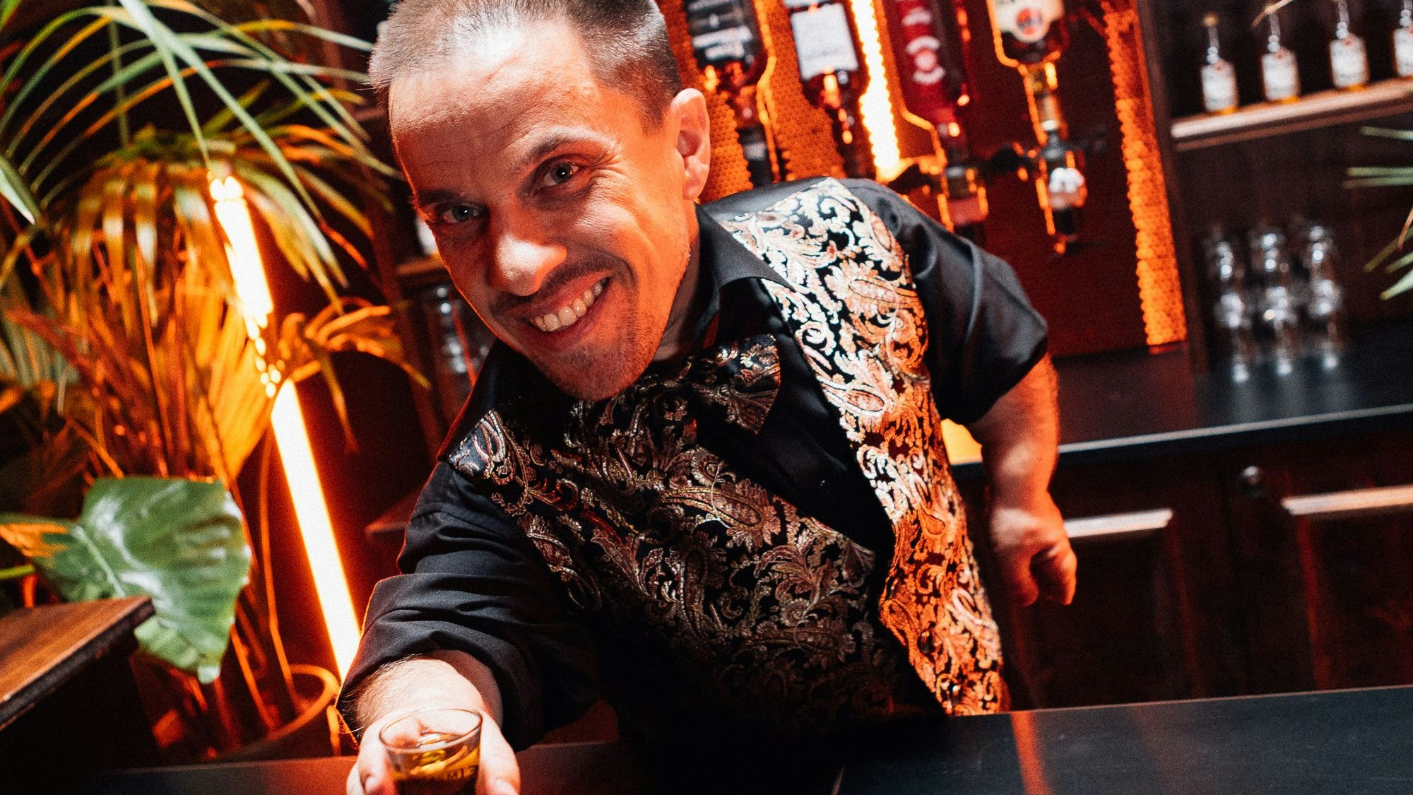 Peter Gatzweiler als Barkeeper serviert Schnaps mit einem Lächeln.
