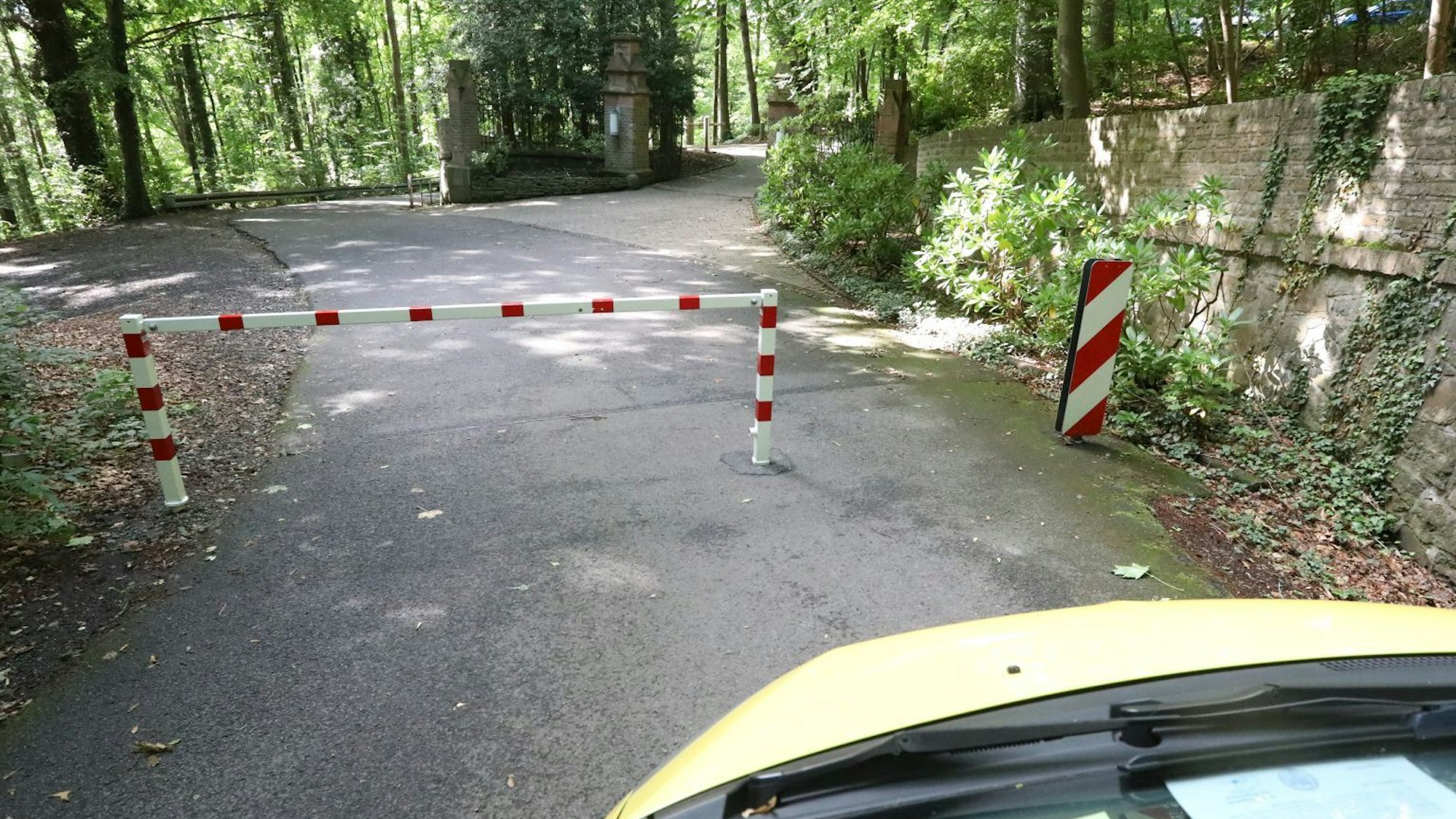 Die Front eines Autos ist im Bild, das vor einer rot-weiß gestreiften Sperre steht. Dahinter ist eine Straße in Wald zu erkennen.