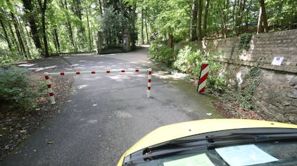 Die Front eines Autos ist im Bild, das vor einer rot-weiß gestreiften Sperre steht. Dahinter ist eine Straße in Wald zu erkennen.&nbsp;&nbsp;
