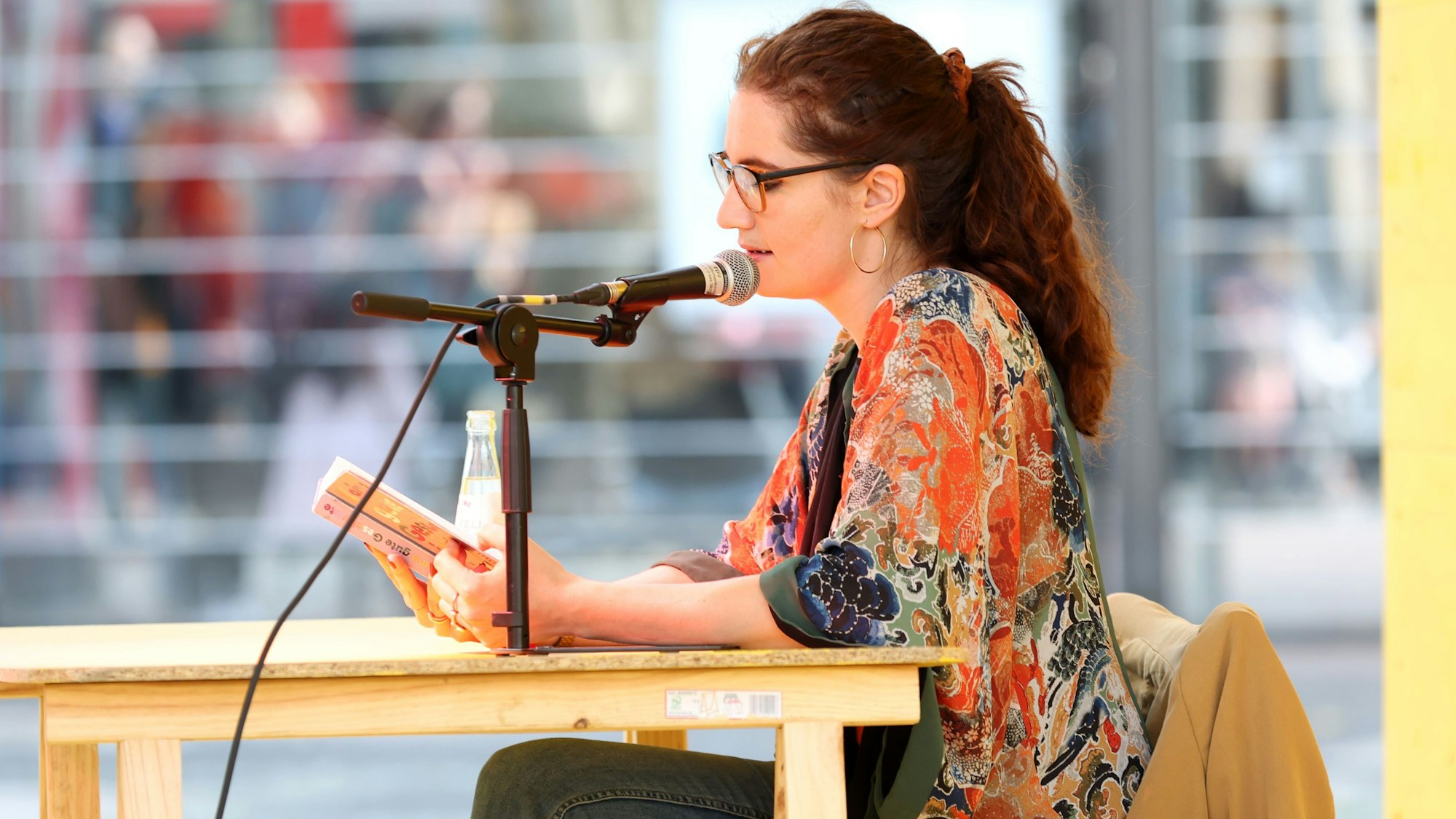 Die Autorin Lisa Roy sitzt mit offenem Buch an einem Tisch und spricht in ein Mikrofon.