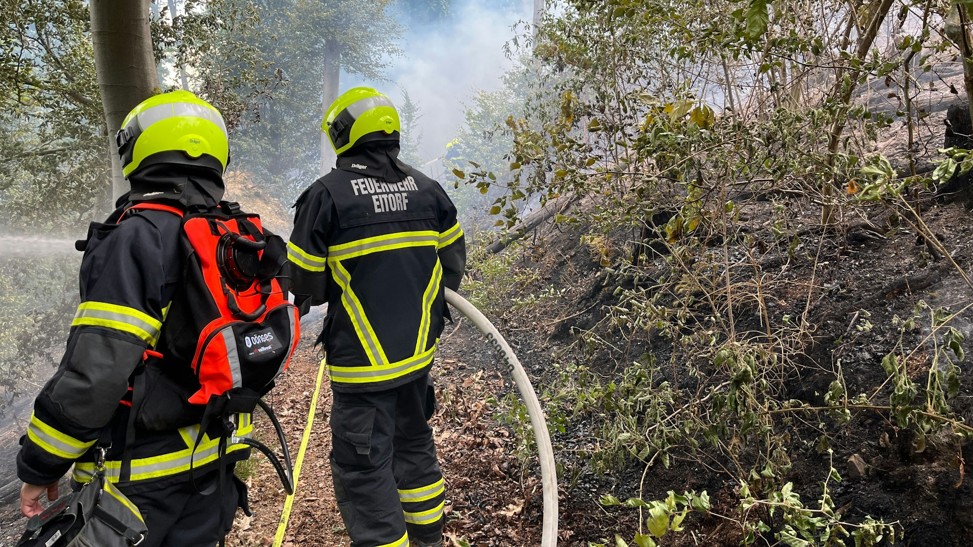 Zwei Einsatzkräfte, die den Schriftzug der Eitorfer Feuerwehr auf dem Rücken tragen, spritzen aus Schläuchen Wasser in den brennenden Wald.