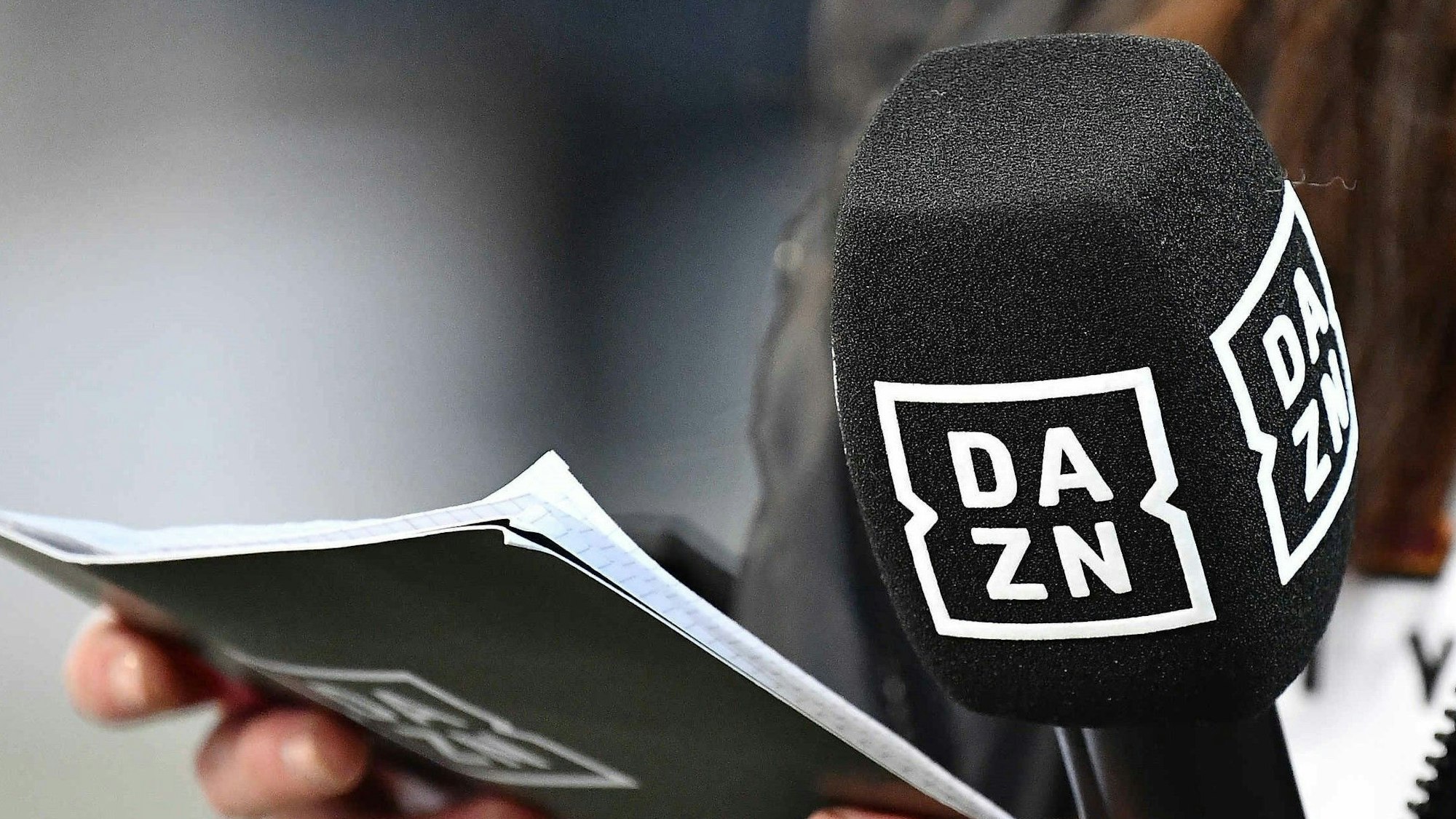Eine Moderatorin hält ein Mikrofon und Moderationskarten mit dem DAZN-Logo in der Hand.