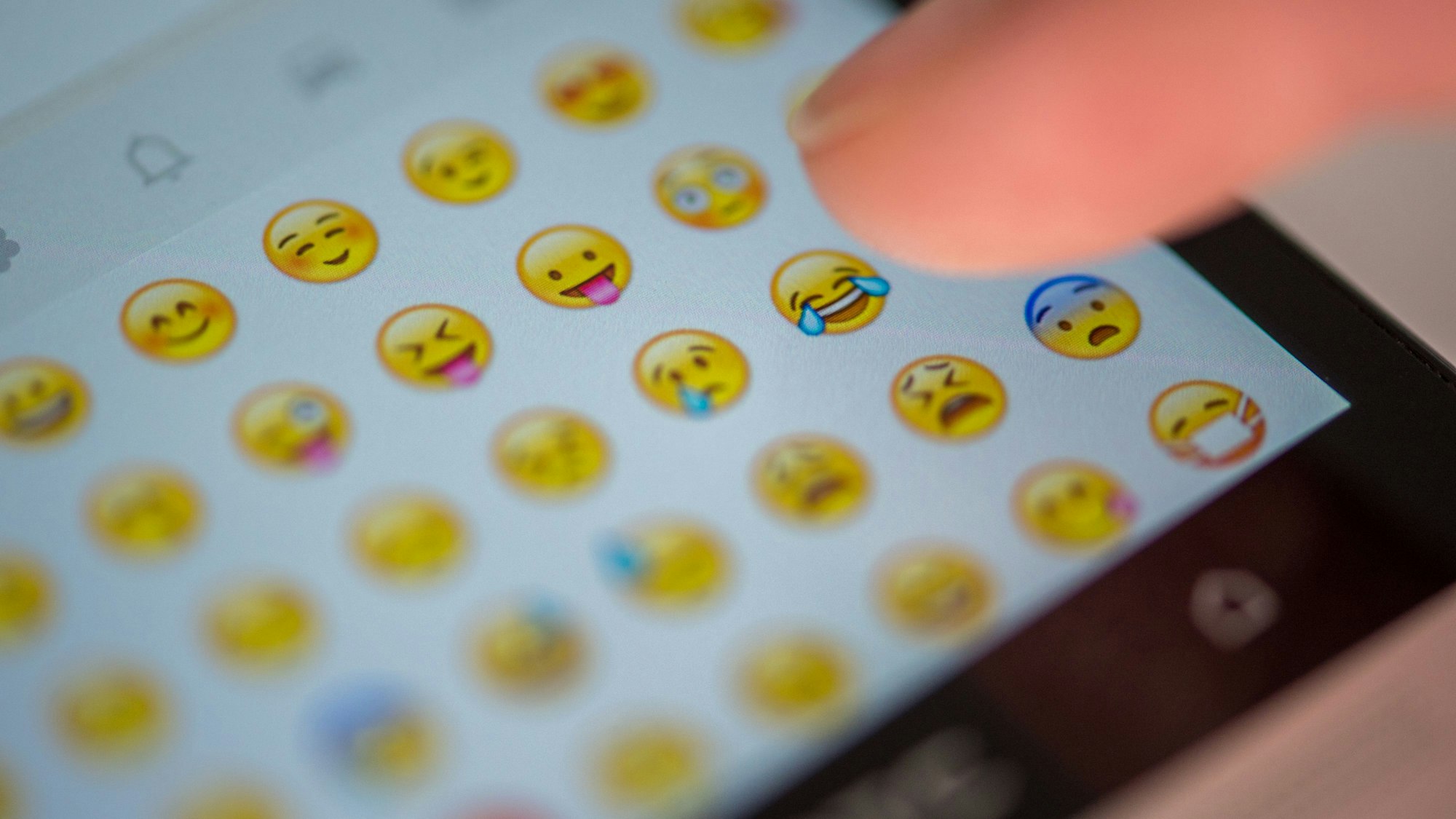 Eine Frau tippt auf das Display eines Smartphones, auf dem zahlreiche Emojis in der Nachrichten-App Whatsapp zu sehen sind.