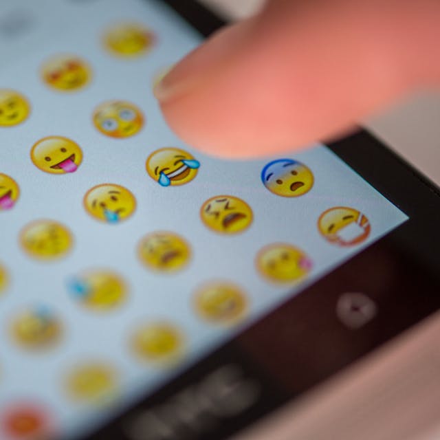 Eine Frau tippt auf das Display eines Smartphones, auf dem zahlreiche Emojis in der Nachrichten-App Whatsapp zu sehen sind.