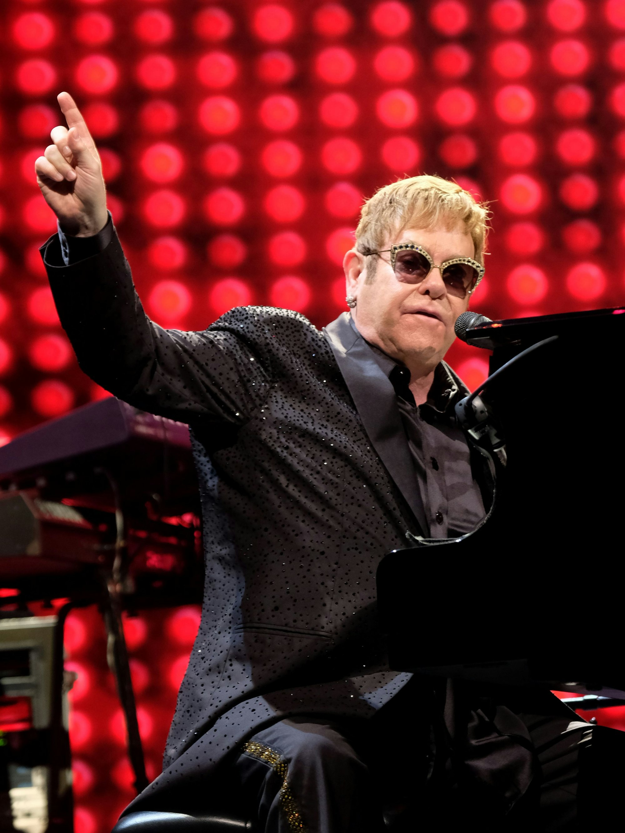 Elton John spielt am Flügel und hebt den Zeigefinger.