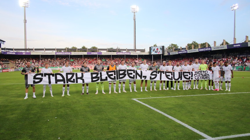 Die Mannschaft von Borussia Mönchengladbach hält ein Banner mit der Aufschrift „Stark bleiben, Stevie!“. Das Foto stammt vom 11. August 2023, im Anschluss an das DFB-Pokalspiel beim TuS Bersenbrück in Osnabrück.