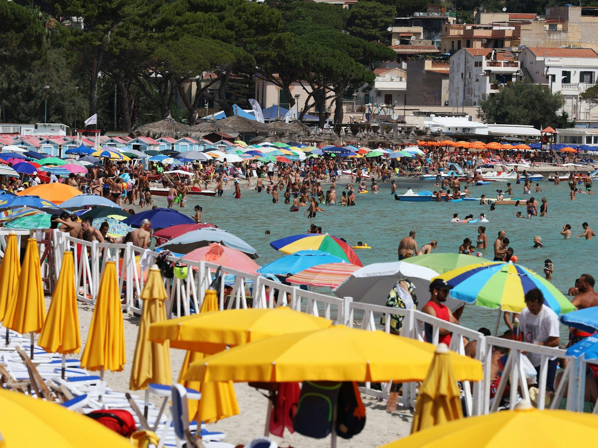 Zahlreiche Menschen sind am Strand in Italien zu sehen. Einige von ihnen baden im Wasser, andere liegen unter gelben Sonnenschirmen.