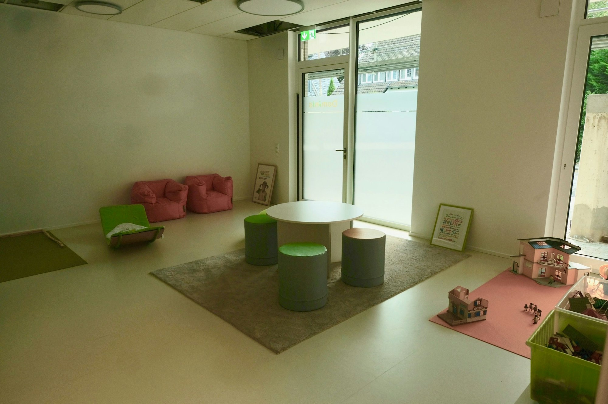 Ein großer heller Raum mit einer Spielecke und einer Sitzgruppe in den Farben grün und rosa ist zu sehen. Der Raum hat bodentiefe Fenster und eine Glastür.
