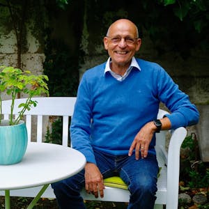 Ein braun gebrannter lachender Mann mit Glatze, Jeans und blauem Pulli sitzt auf einer weißen Bank an einem weißen Tisch, auf dem eine üppige Grünpflanze steht.