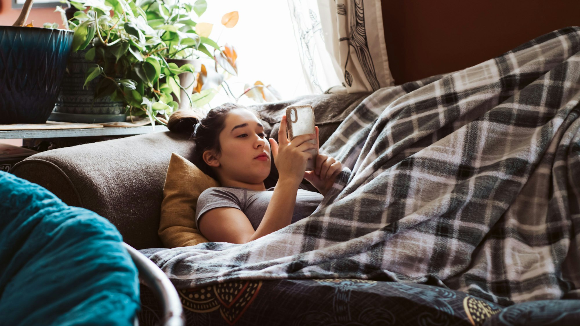 Eine Jugendliche, die unter einer Decke auf der Couch liegt und etwas auf dem Handy sieht.