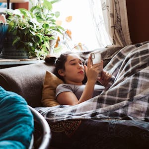 Eine Jugendliche, die unter einer Decke auf der Couch liegt und etwas auf dem Handy sieht.