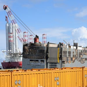 Der schwer beschädigte Frachter „Fremantle Highway“ liegt im Hafen von Eemshaven an der niederländischen Nordseeküste. Sie wird von einem Notfallschlepper gestützt. Davor sind gelbe Container aufgebaut.