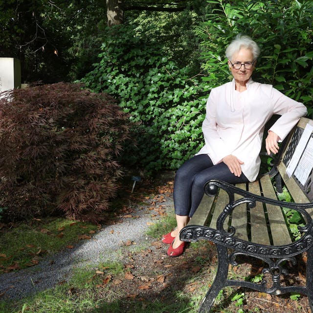 Dorothee Fiedler sitzt auf einer Bank aus Holz mit einem Gestell aus Metall. Neben ihr ist das Grab ihres Sohnes.