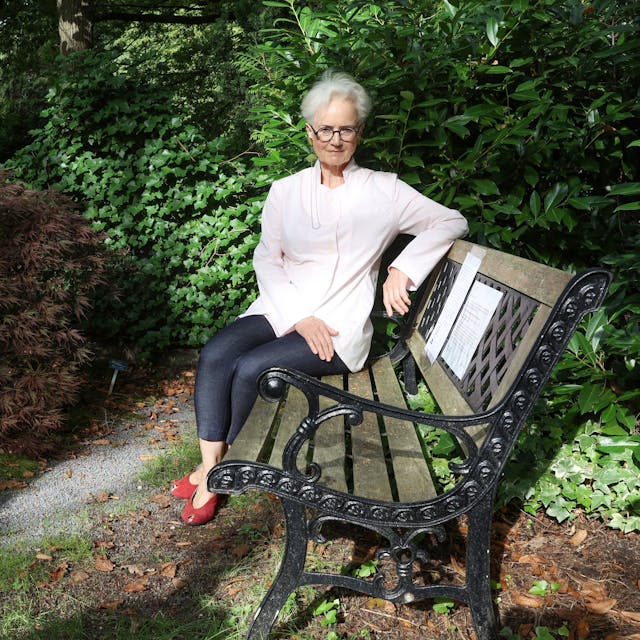 Dorothee Fiedler sitzt auf einer Bank aus Holz mit einem Gestell aus Metall. Neben ihr ist das Grab ihres Sohnes.